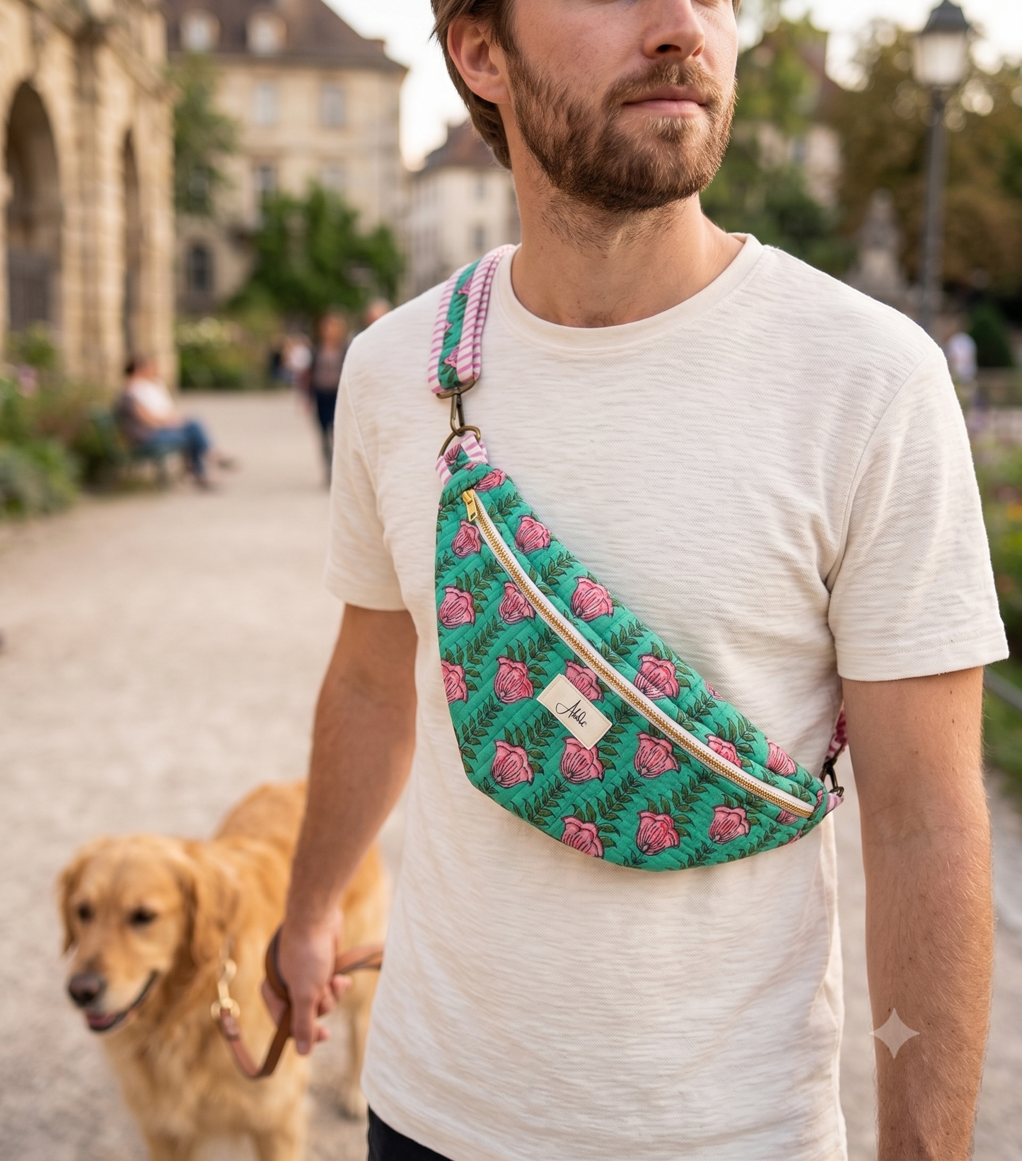 A man wearing a cream-colored t-shirt walks a golden retriever dog in a park while wearing a green floral crosstrain sling bag with pink roses and a beige zipper.