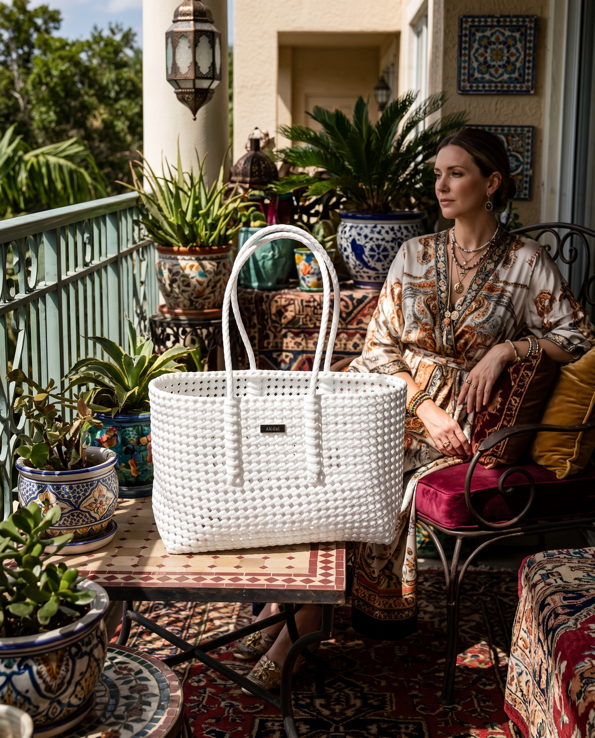 A woman sits on a wrought-iron chair on a decorated balcony, wearing a patterned dress and layered necklaces, while a large white woven recycled plastic tote bag sits on a small table in the foreground. The setting features potted plants, patterned ceramic pots, and a red patterned rug.