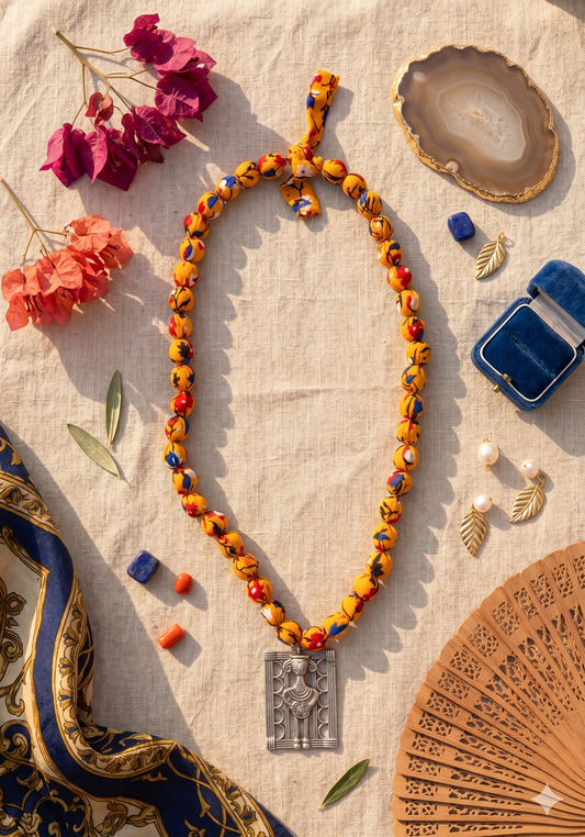 A colourful deep yellow beaded necklace with a Mettalic pendant on a textured surface with decorative items.