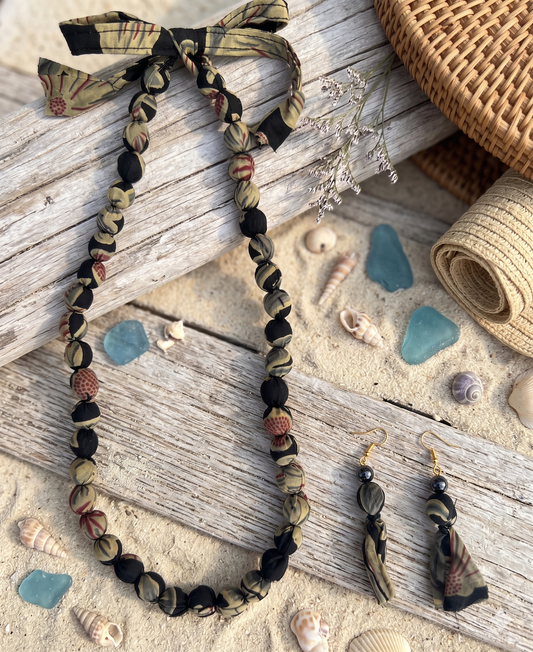 A fabric necklace in black & beige colour with matching earrings with a beach-themed design on a wooden surface with shells and stones.