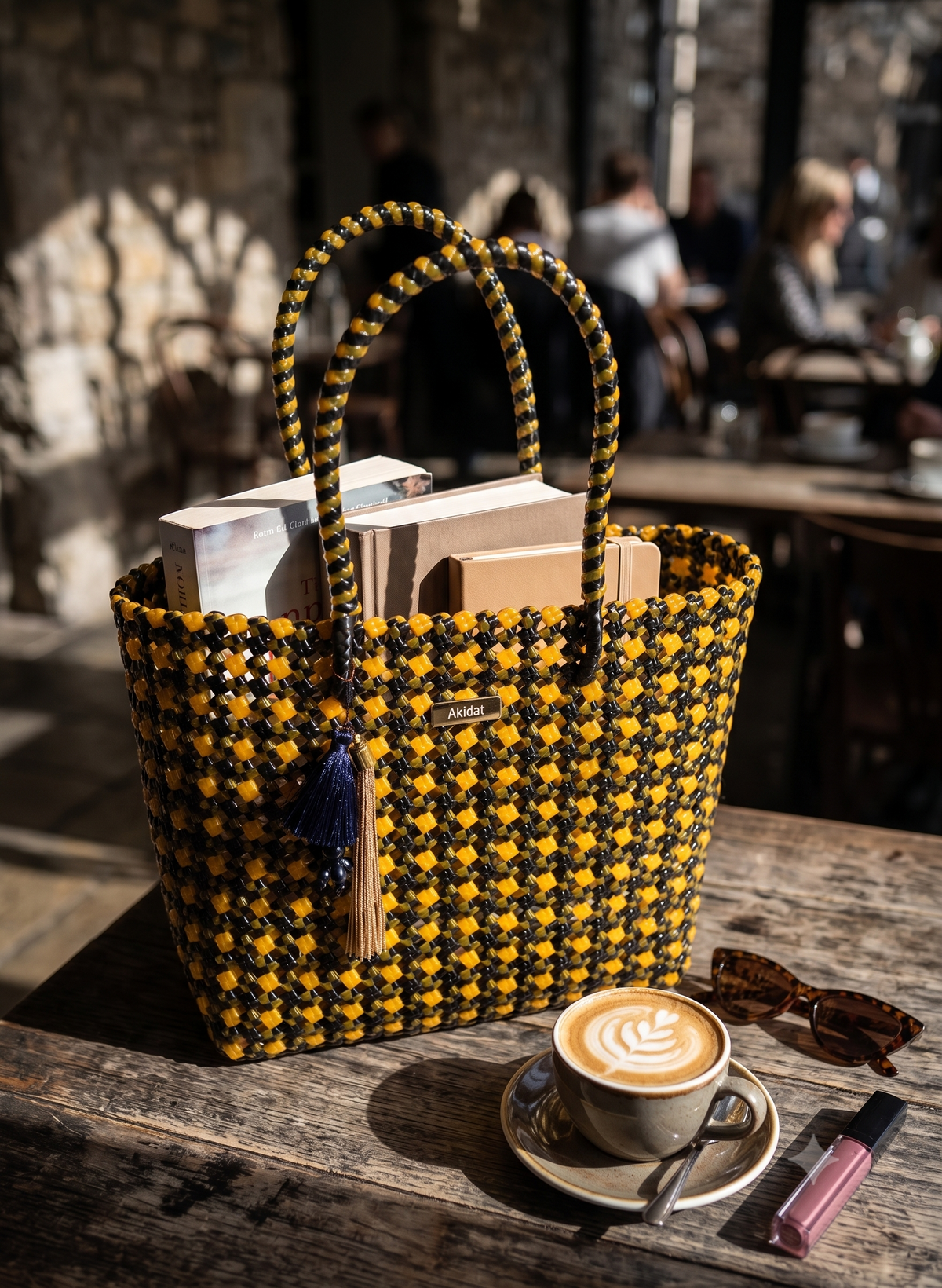 A woven recycled plastic tote bag in black and yellow sits on a rustic wooden table in a cafe setting, filled with books and magazines. A cup of latte, sunglasses, and a lip product rest beside it.