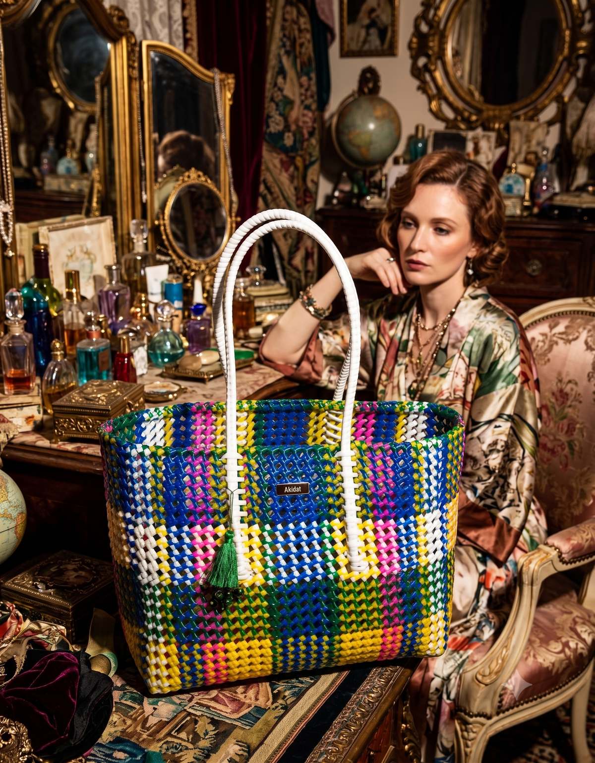 A woman in a patterned satin robe sits in an ornate vintage room, leaning towards a large woven recycled plastic tote bag with a colourful plaid pattern and white handles.