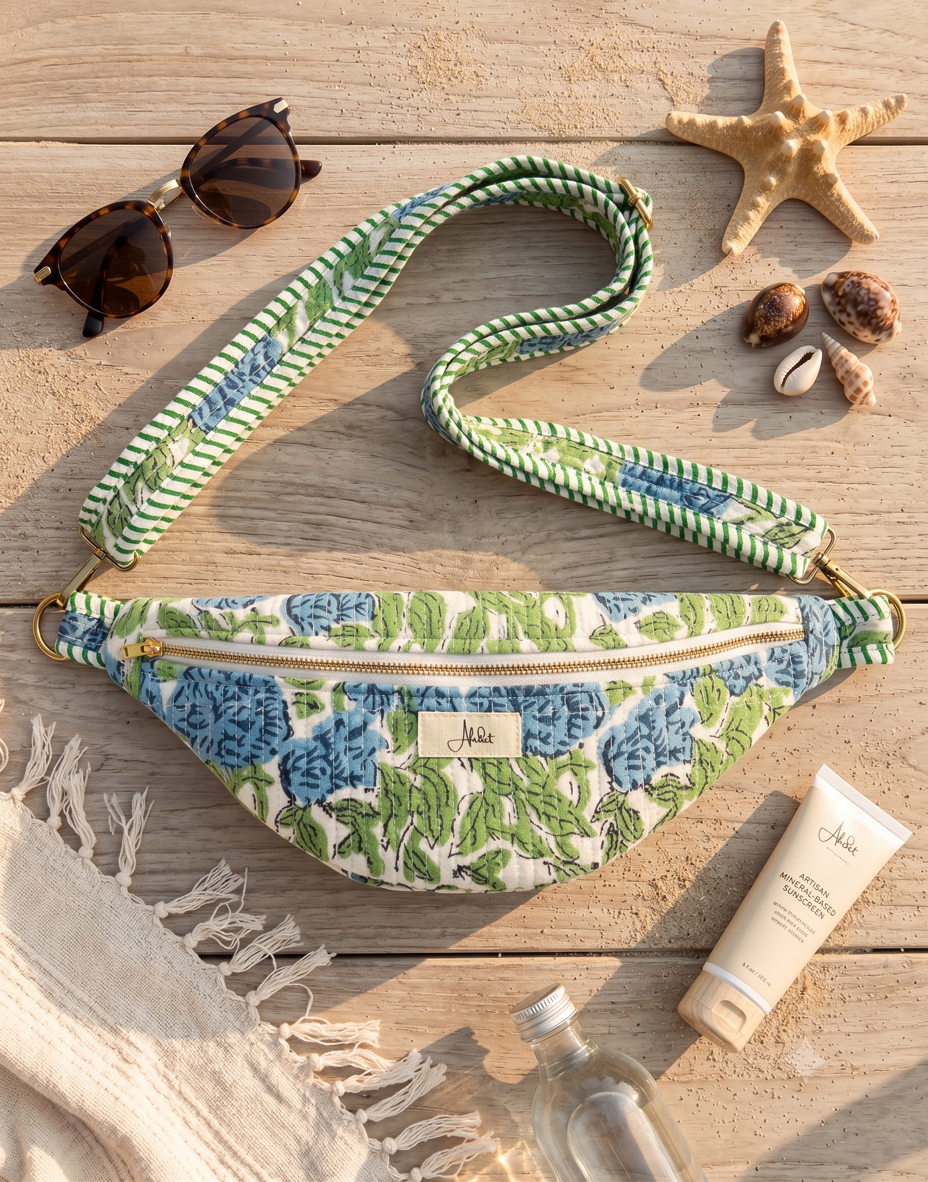 A floral fanny pack with a green and white striped woven strap, styled on a wooden surface with beach-themed props including sunglasses, a starfish, seashells, a tube of lotion, and a bottle.