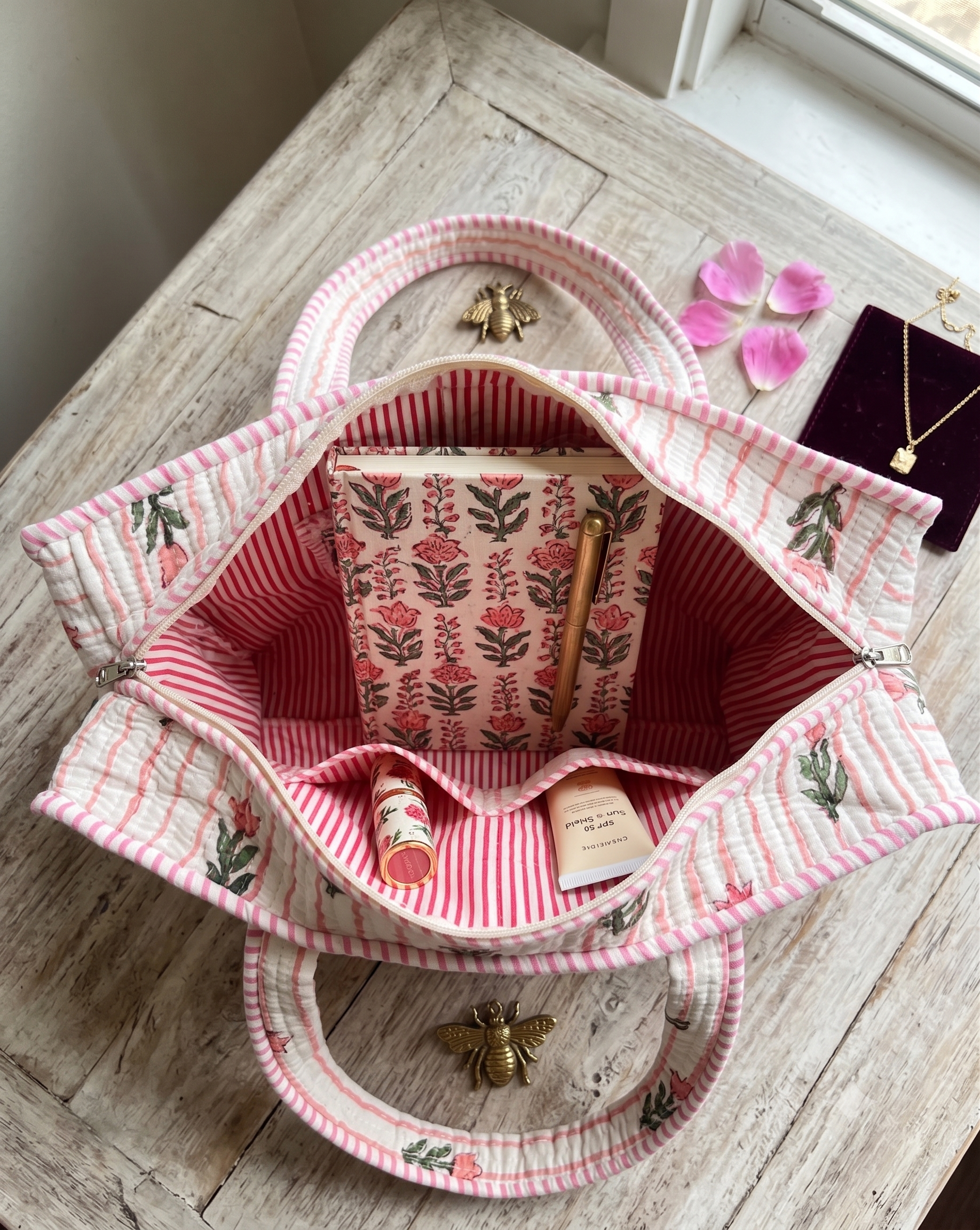 A pink and white striped quilted tote bag with floral prints with white and pink striped interiors carrying a notebook, pen, lipstick, and SPF, shown open on a rustic wooden table.