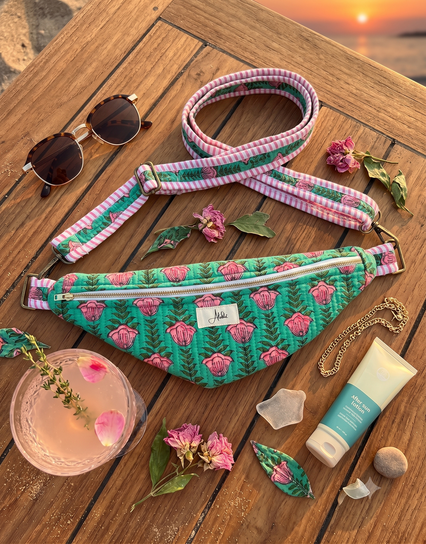 A green and pink floral cross-body bum bag on a wooden table alongside sunglasses, a necklace, body lotion, a cold drink and some dried flowers. 
