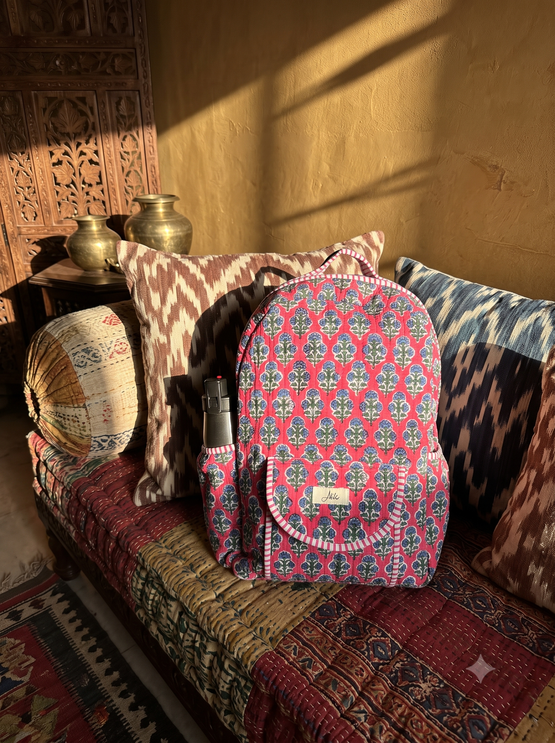 A pink patterned backpack with a floral Blockprint sits on a patterned sofa in a sunlit room with traditional decor, including pillows, a carved wooden cabinet, and brass vases.