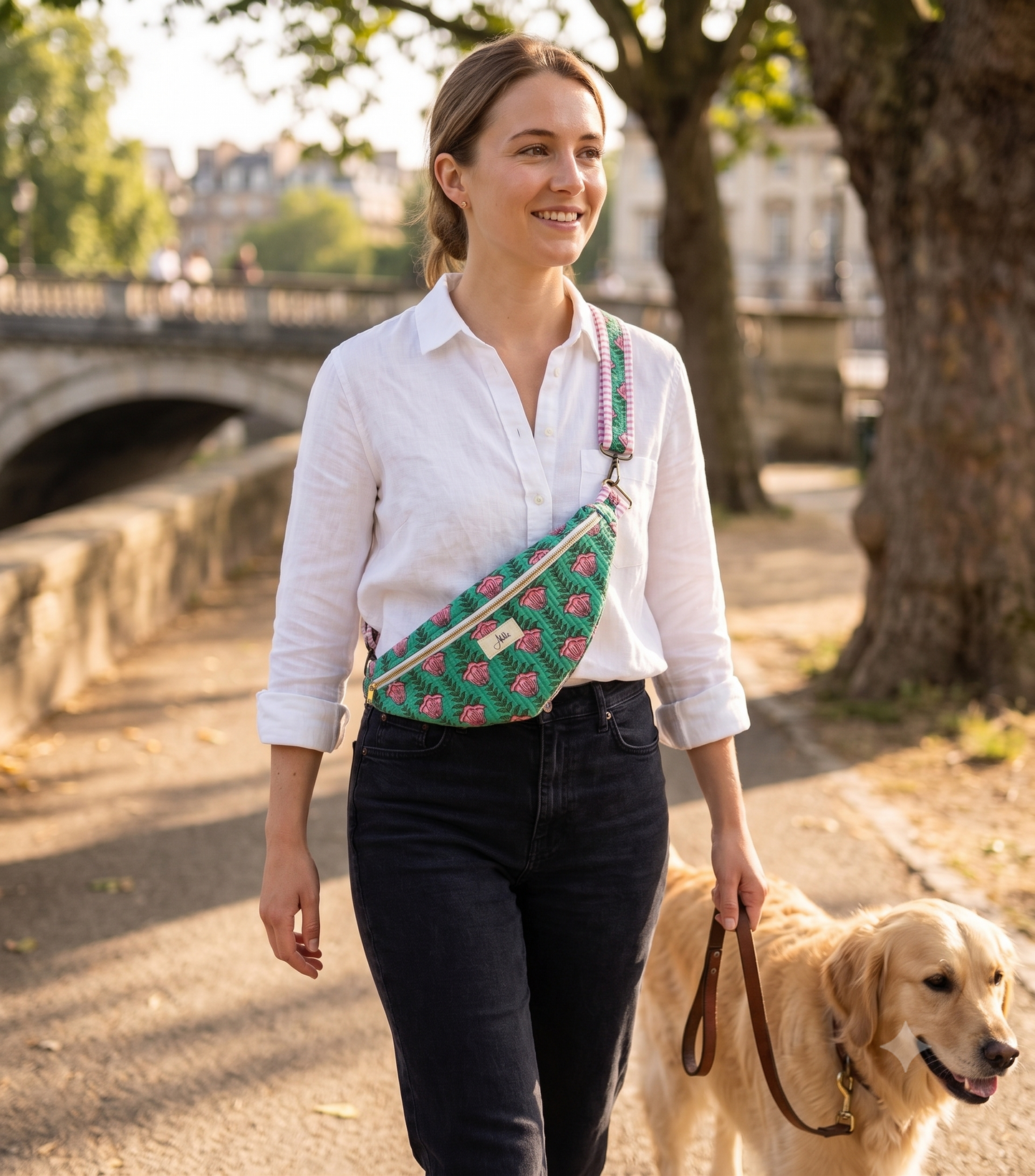 A woman wearing a white shirt and black jeans is walking a golden retriever, with a green and pink floral cross-body fanny bag, next to a canal 