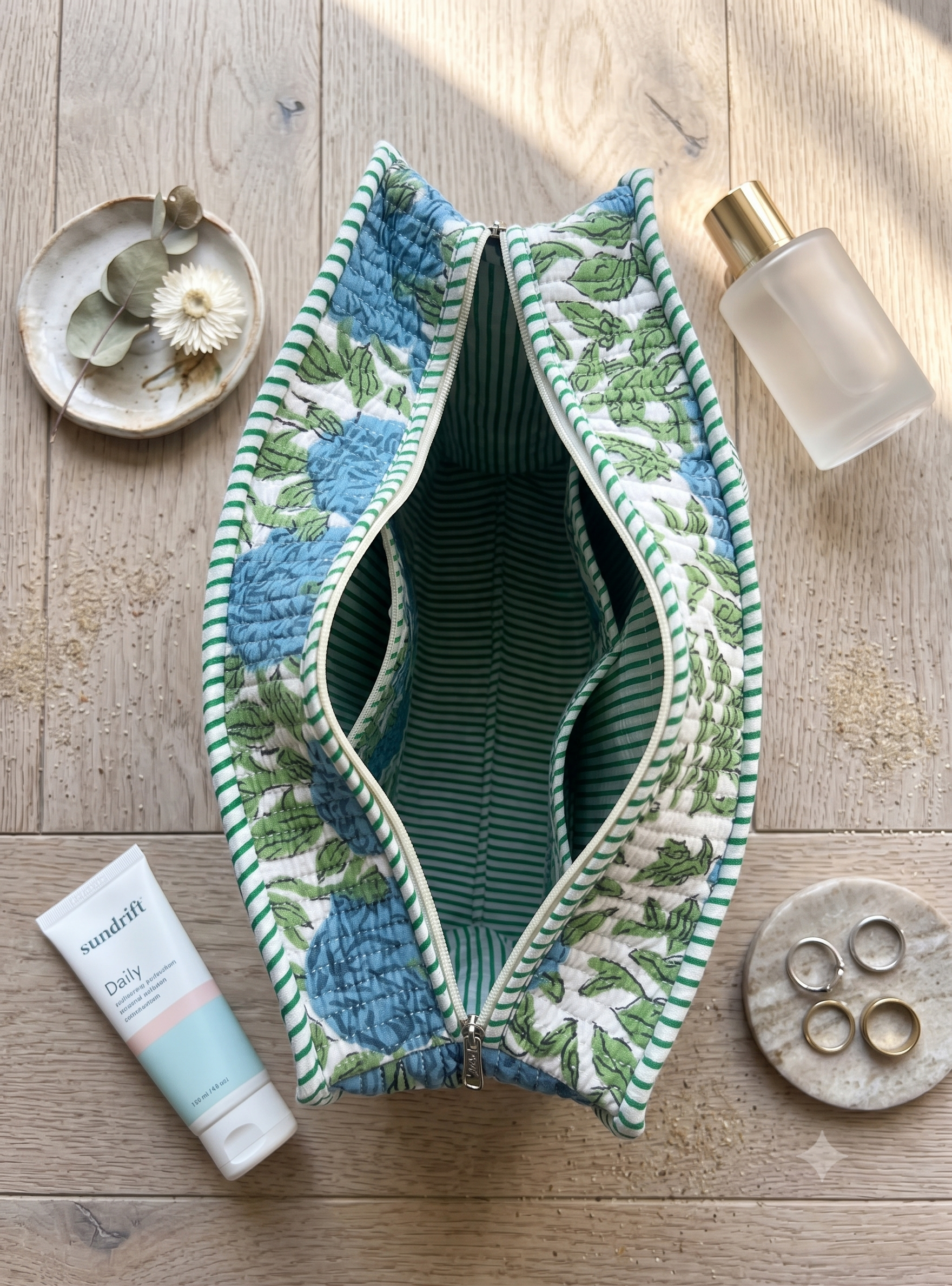 A quilted tote bag with a blue and green floral pattern and striped green interior, laid flat on a light wood surface with styled props including a tube of sunscreen, a bottle, rings, and a small dish. The inside of the bag showcases 3 pockets. 