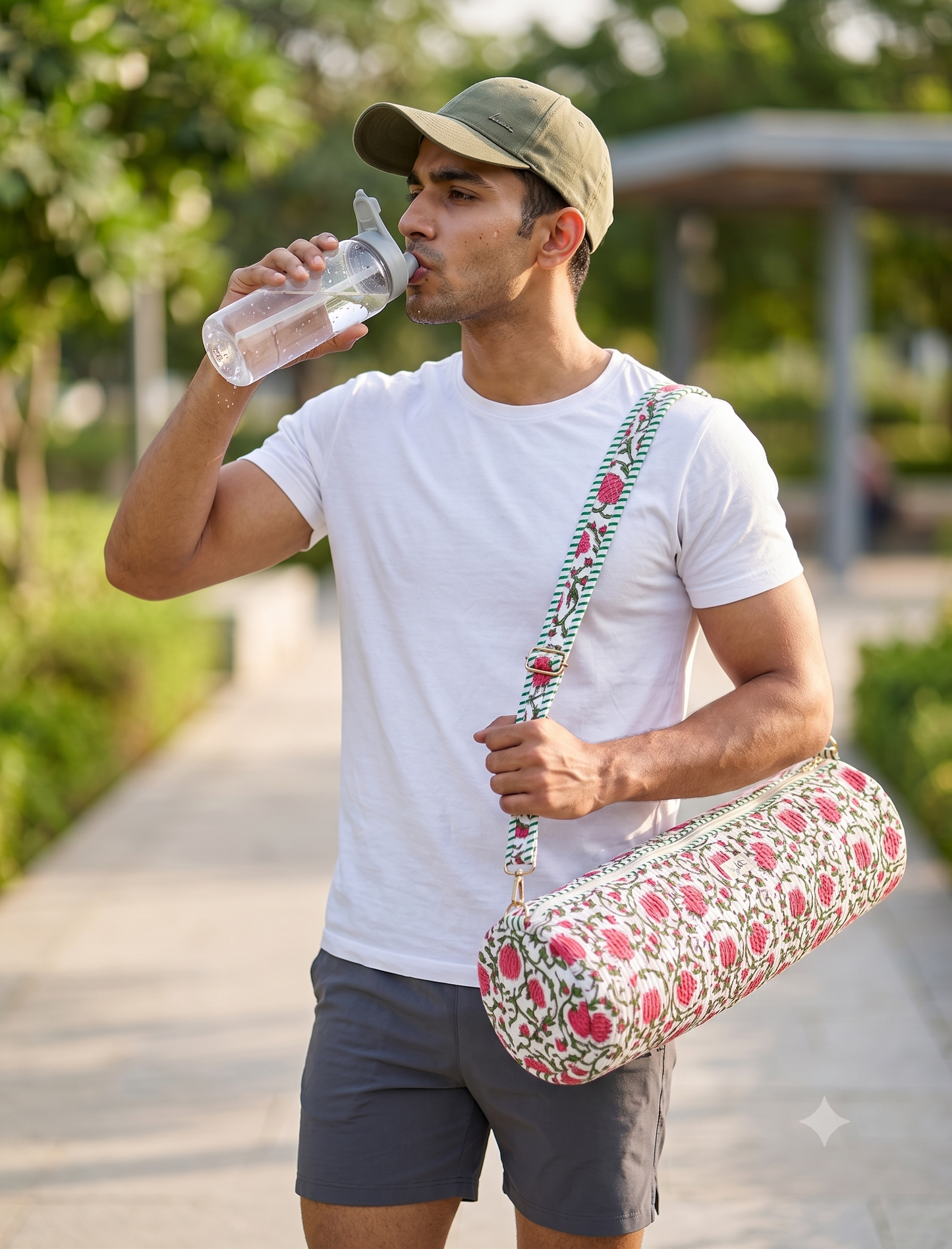 A man in a white t-shirt and gray shorts walking outdoors on a park path while drinking from a clear water bottle. He is wearing a green cap and carrying a floral patterned yoga mat bag with a pink and green floral print and a matching patterned shoulder strap.