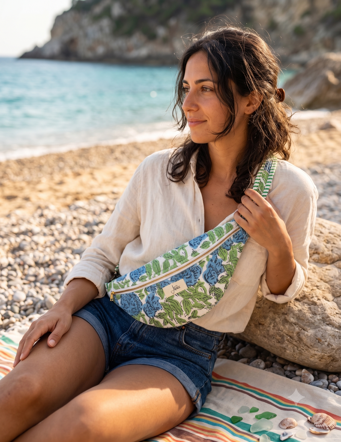 A woman sitting on a pebble beach wearing a cream shirt and denim shorts, carrying a green and blue floral-patterned crossbody fanny bag.