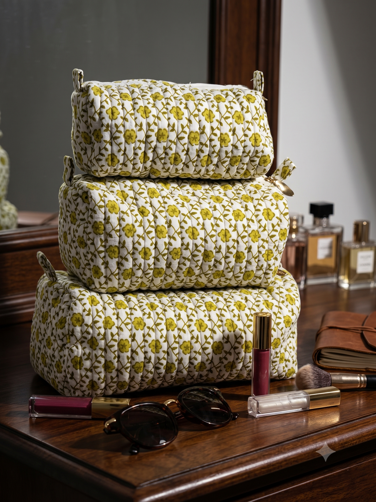 Set of 3 vanity kits/toiletry bags in different sizes sitting on a wooden dressing table with makeup products and sunglasses. 