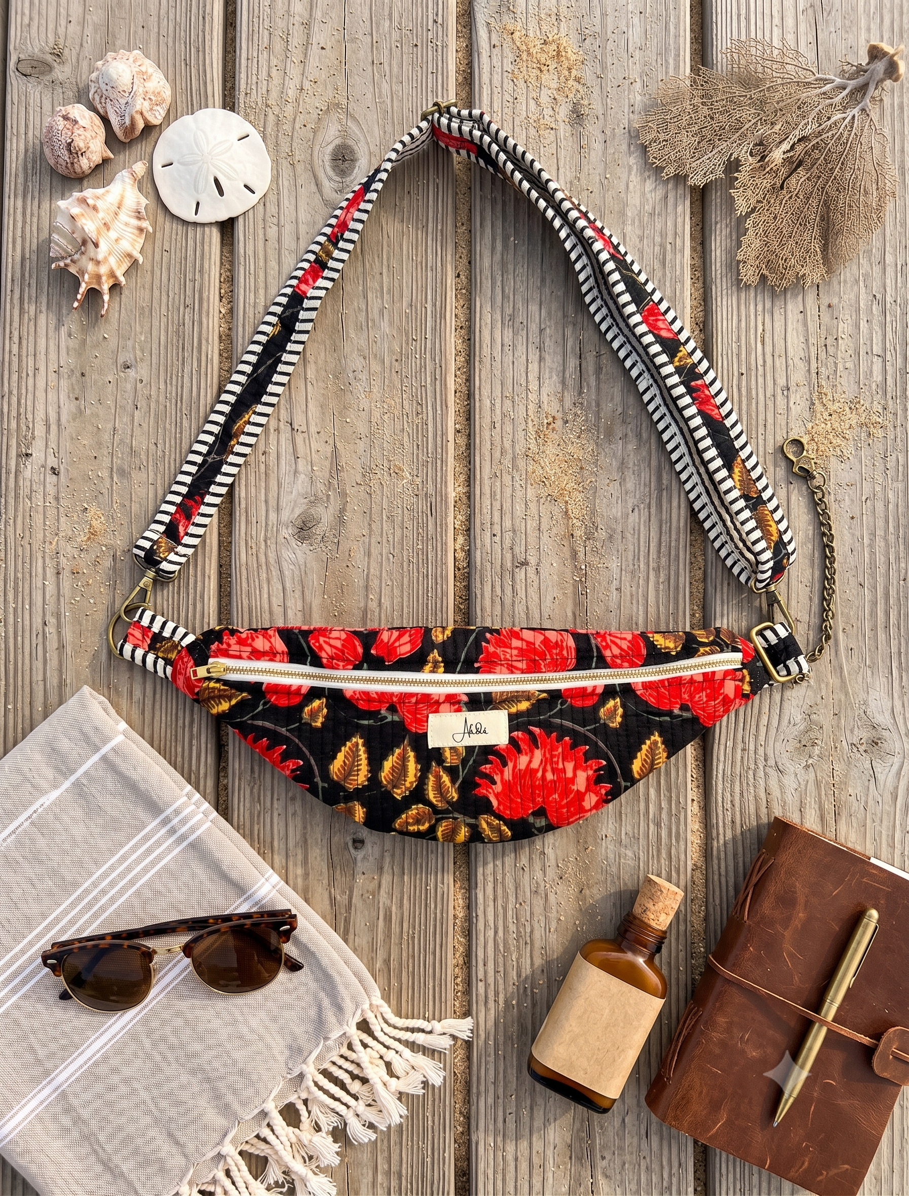 A black and red floral fanny pack with a wide patterned strap, styled in a flat lay on a rustic wooden background with beach-themed props including seashells, sunglasses, a notebook, and a bottle.