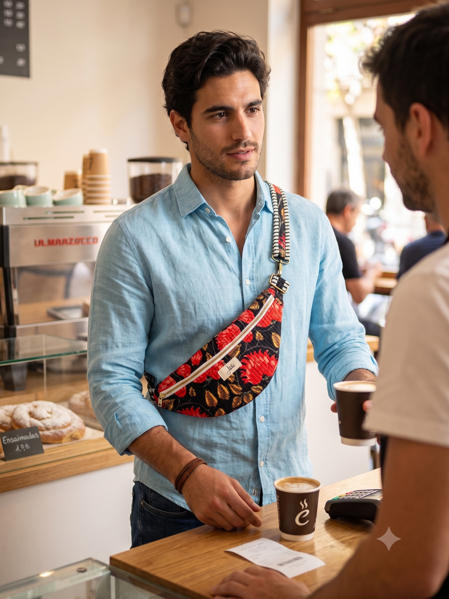 A man in a light blue shirt wearing a red, black, and gold patterned crossbody fanny pack while standing at a coffee shop counter.