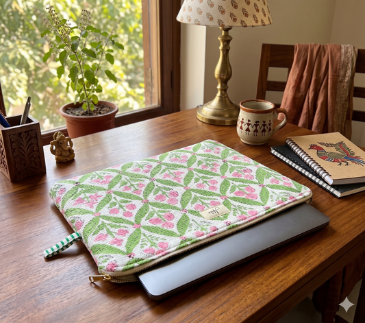 Quilted laptop sleeve with green and pink floral block print pattern on white base on a wooden table with a brass lamp, coffee mug and notebooks.