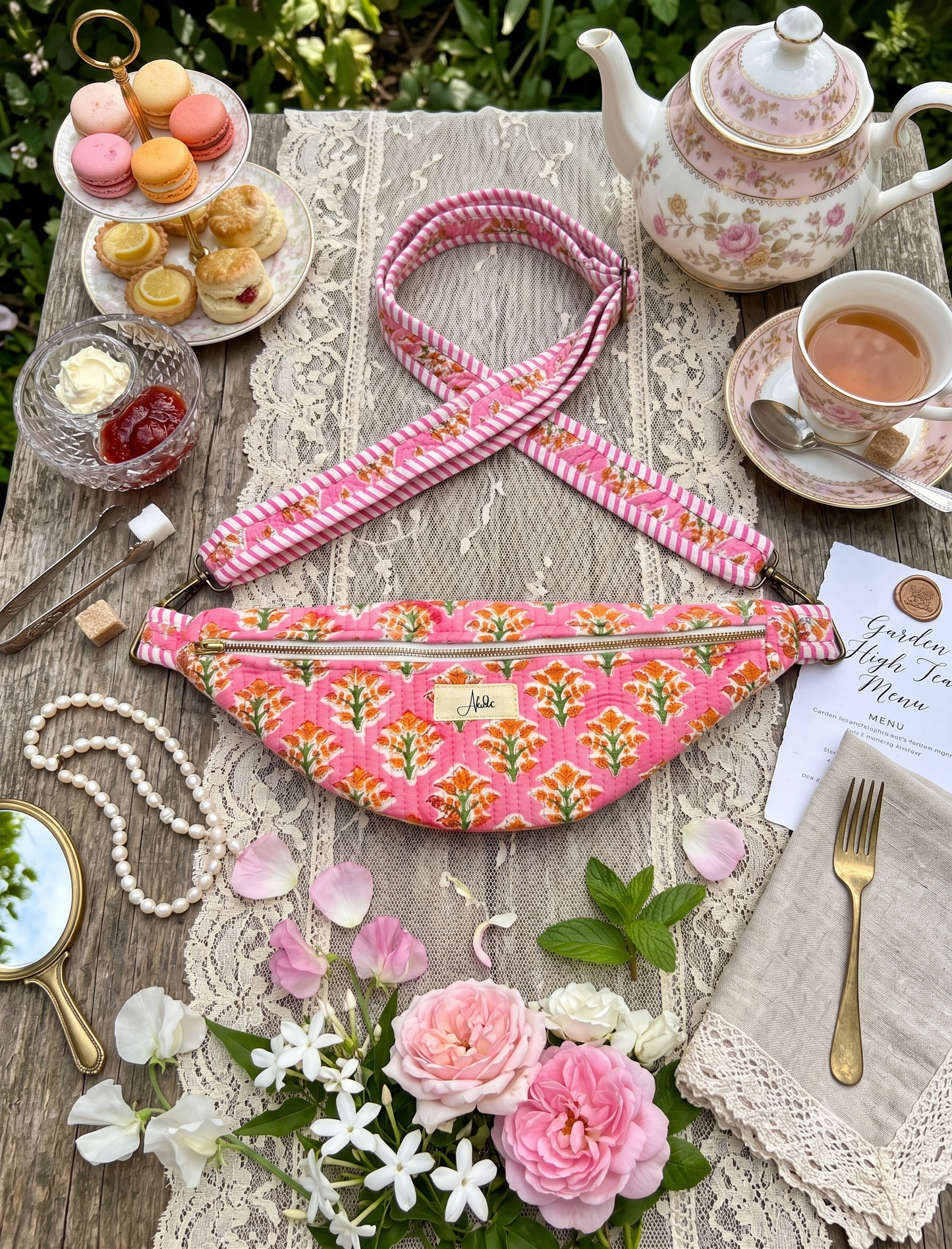 A pink floral fanny pack with a striped shoulder strap is styled in a tea-time setting on a lace table runner, surrounded by a teapot, cup of tea, macarons, and flowers.