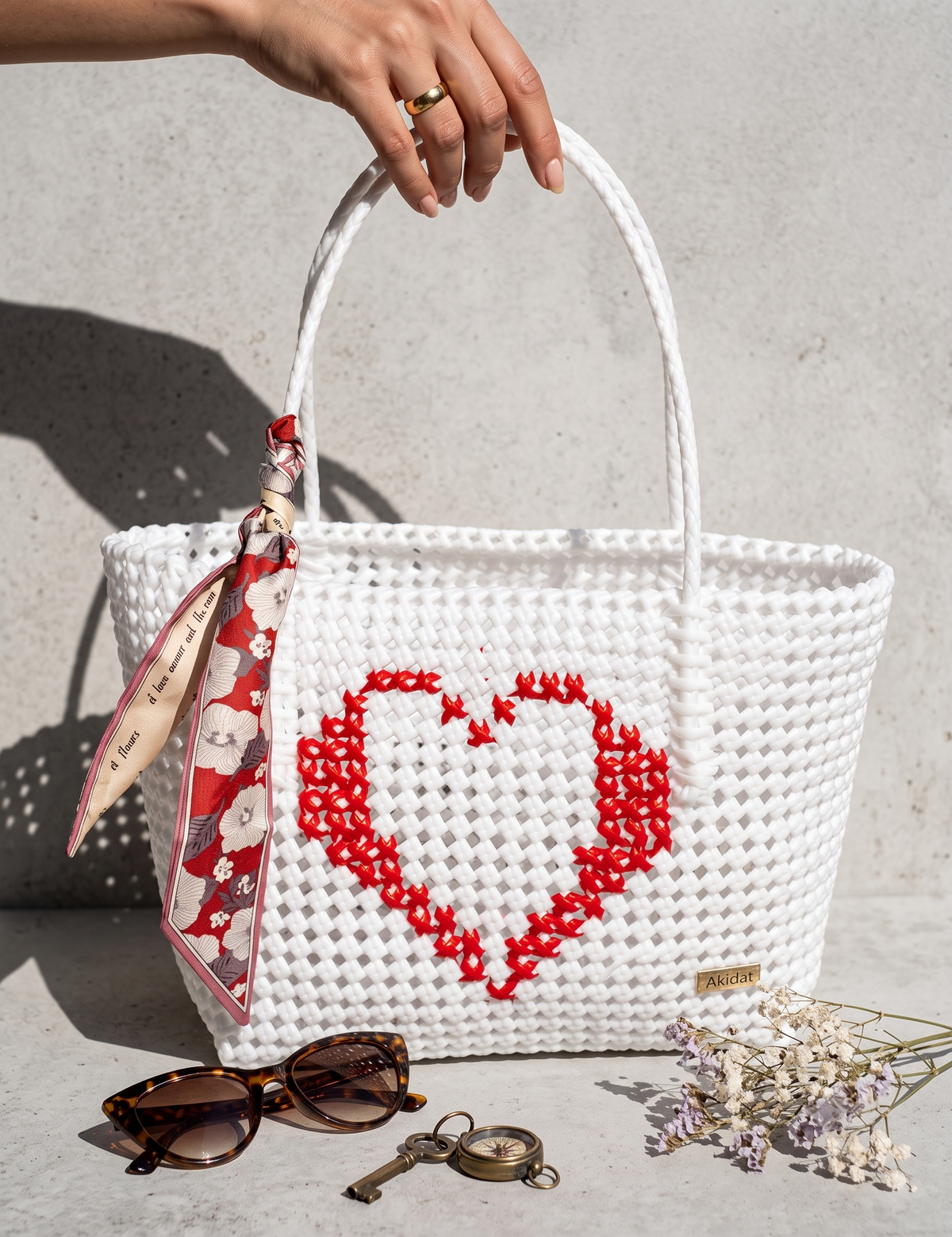 A white woven recycled plastic tote bag with a red heart design on the front, held by a hand. A patterned scarf is tied to the handle, and the bag is styled with sunglasses, a vintage key, and dried flowers on a concrete surface.