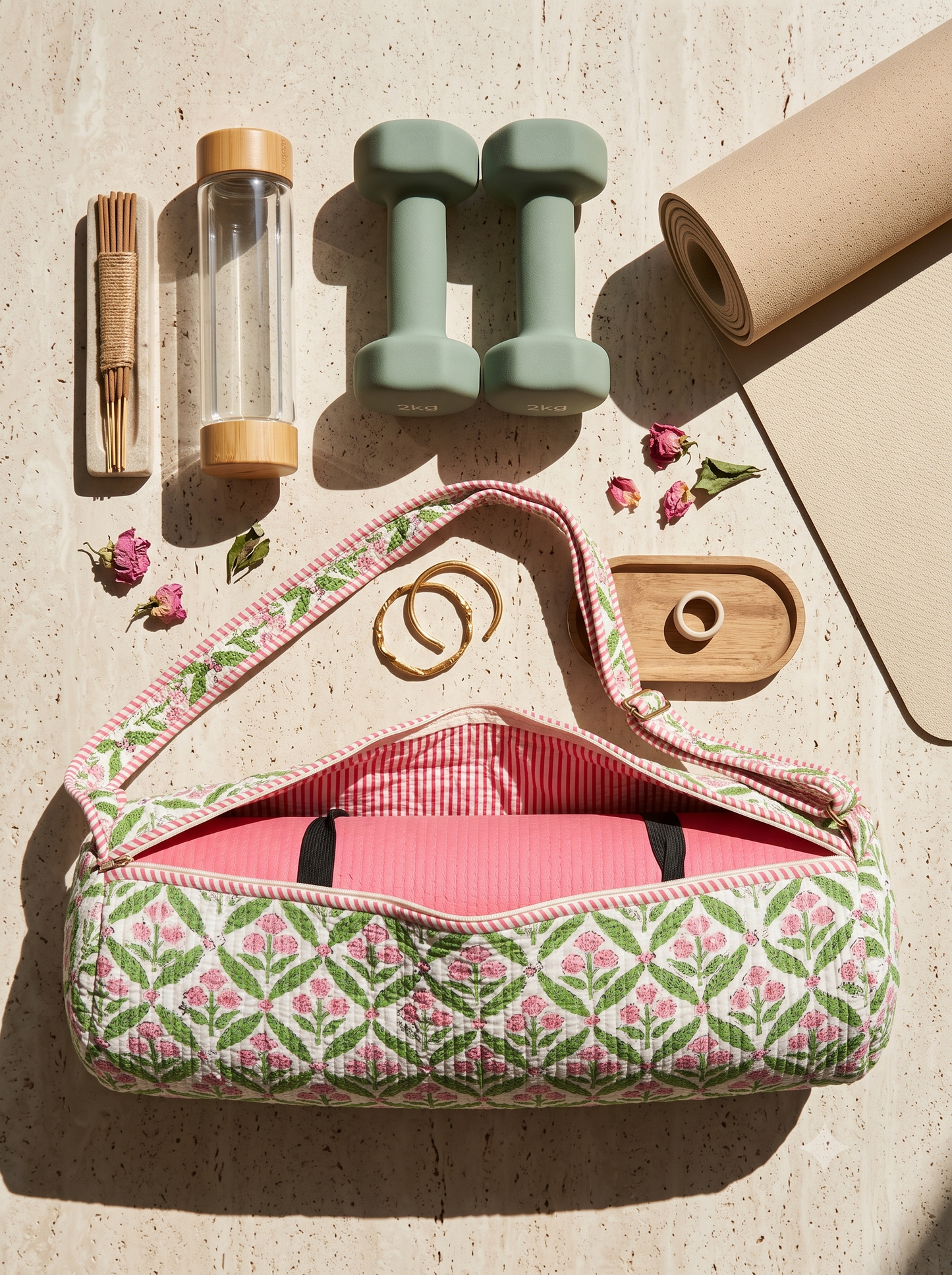 A quilted yoga carry bag with a pink and green floral pattern, shown open with a pink yoga mat inside. The bag is styled in a flat-lay arrangement with a beige yoga mat, two mint green dumbbells, a glass water bottle, a wooden incense holder, and small dried flowers on a light beige textured surface.