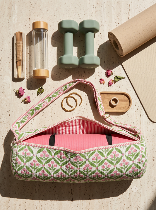 A quilted yoga carry bag with a pink and green floral pattern, shown open with a pink yoga mat inside. The bag is styled in a flat-lay arrangement with a beige yoga mat, two mint green dumbbells, a glass water bottle, a wooden incense holder, and small dried flowers on a light beige textured surface.