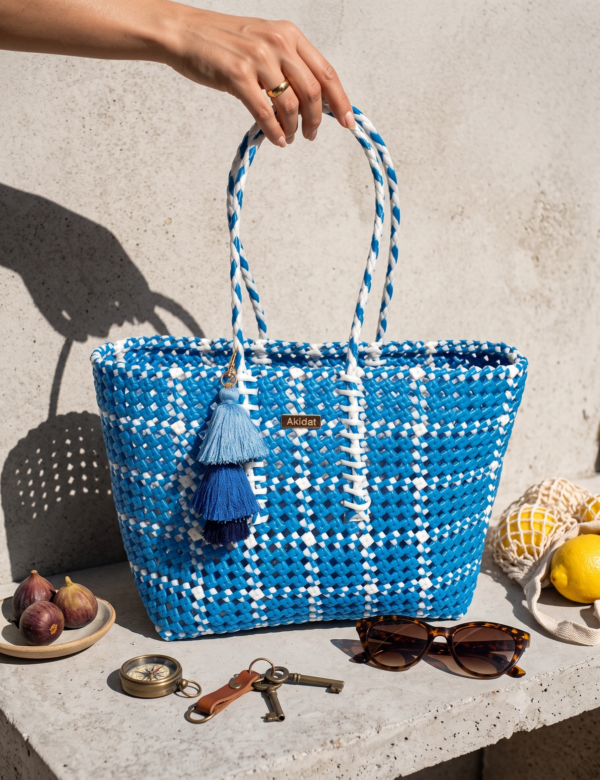 A blue and white woven recycled plastic tote bag with braided handles and tassel charms, held by a hand against a textured concrete wall. The bag is styled on a stone ledge with props including sunglasses, keys, figs, and lemons.