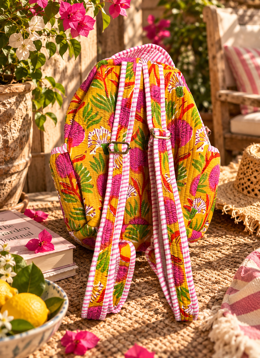 A colourful backpack with a tropical floral print in yellow, pink, green, and orange, featuring pink-and-white striped adjustable shoulder straps, set in a warm, sunlit outdoor patio scene.