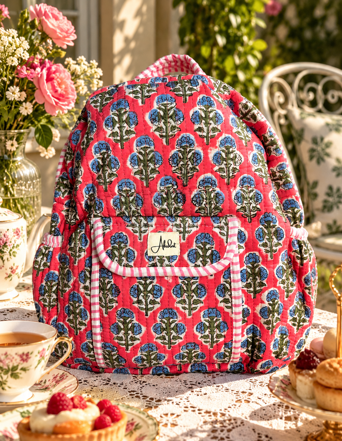 A quilted fabric backpack with a pink, blue, and green floral damask pattern and striped trim, styled on an outdoor patio table with a teacup, pastries, and flowers.