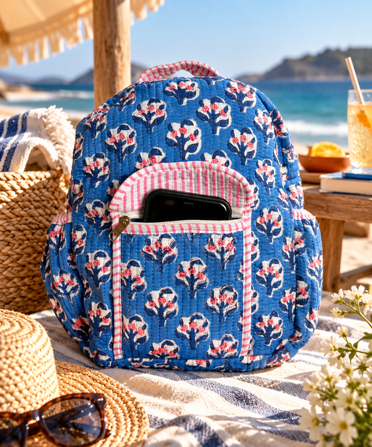A blue quilted backpack with a pink and white floral pattern and a front zipper pocket with a smartphone inside, sits on a beach blanket at the beach, with sunglasses, a straw hat, and a drink nearby.