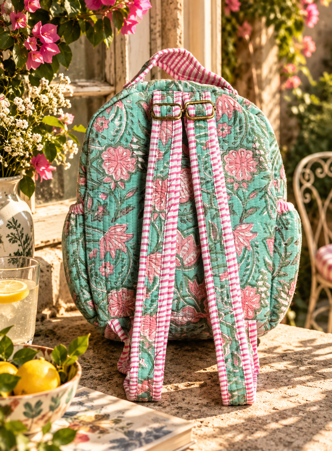 A quilted fabric backpack with a green and pink floral pattern and pink-and-white gingham adjustable straps, displayed on a stone table in a sunlit garden setting.