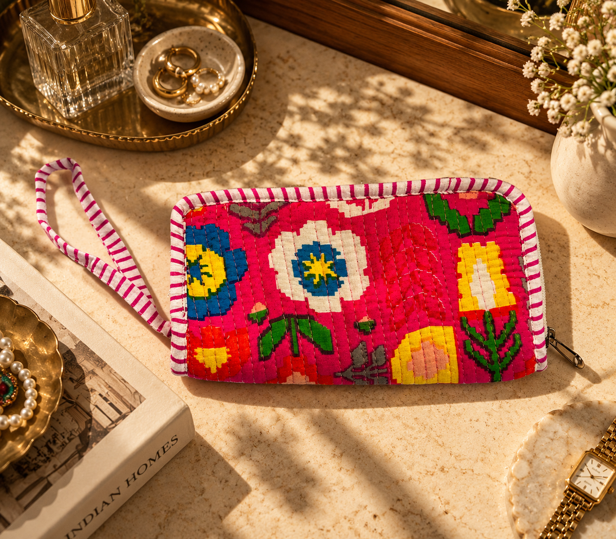 Pink-coloured cotton quilted zippered wallet with a hand string on the side to hold lying on a marble table with other accessories. 