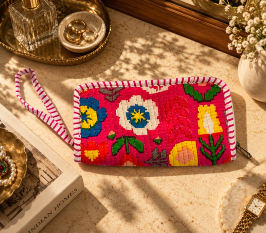 Pink-coloured cotton quilted zippered wallet with a hand string on the side to hold lying on a marble table with other accessories. 