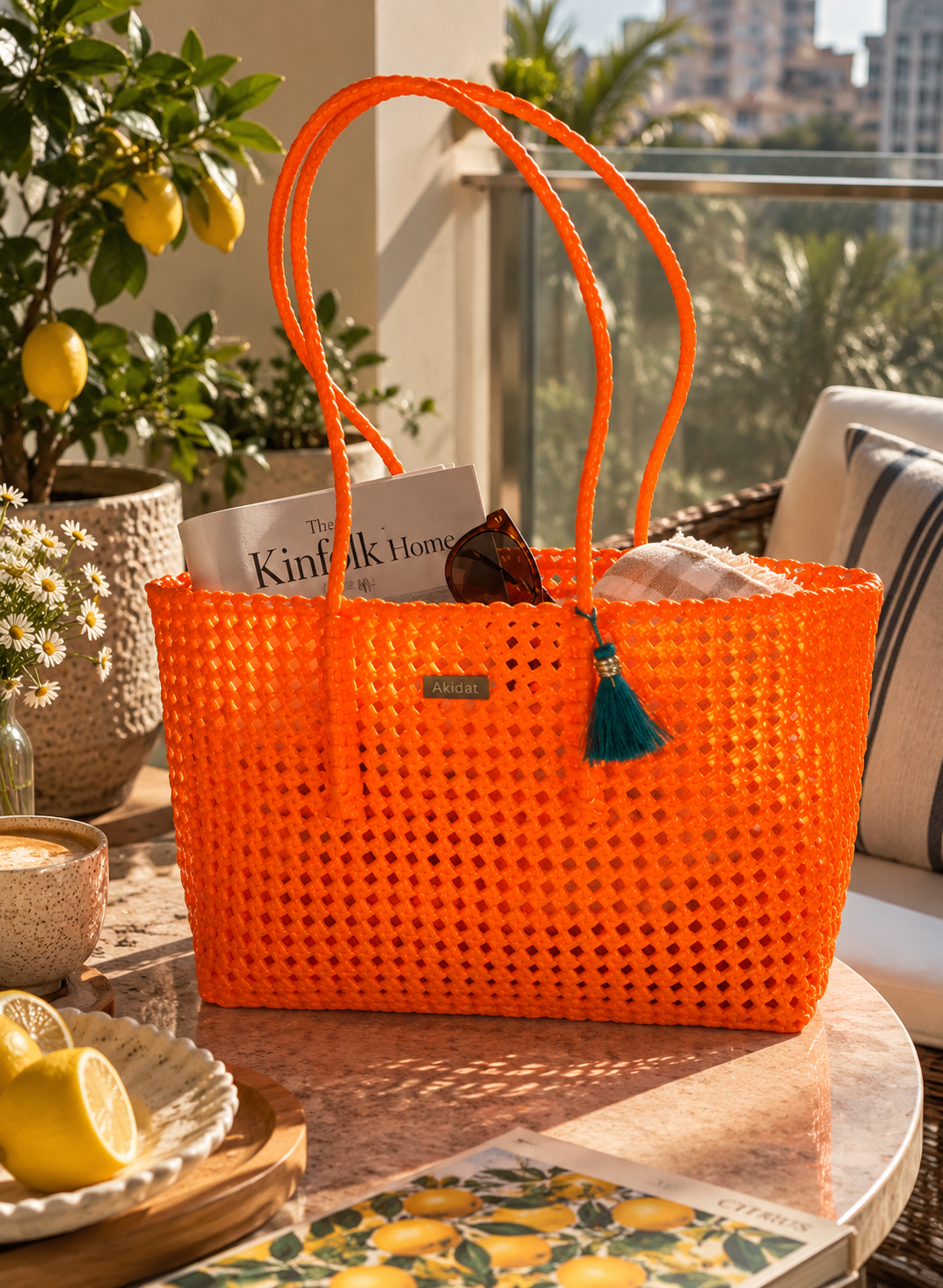 Bright orange woven recycled plastic tote bag with double shoulder straps, placed on a round table in a sunlit outdoor patio setting with lemon plants, a cushion, and a magazine visible inside.
