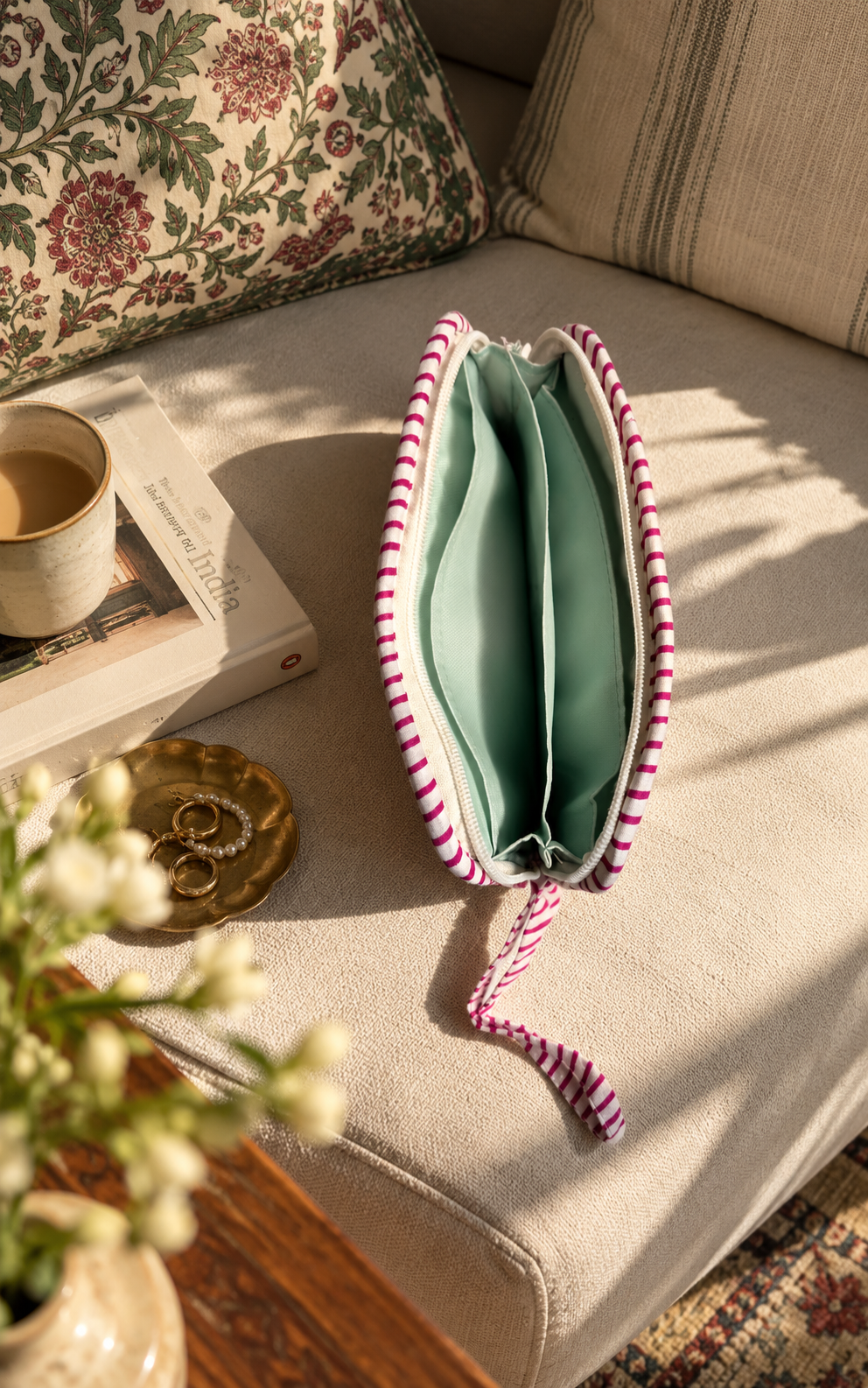 A zippered pouch with a pink and white striped fabric exterior and a water-resistant interior, resting on a beige sofa in a sunlit room with decorative pillows, a book, a cup, and flowers nearby.