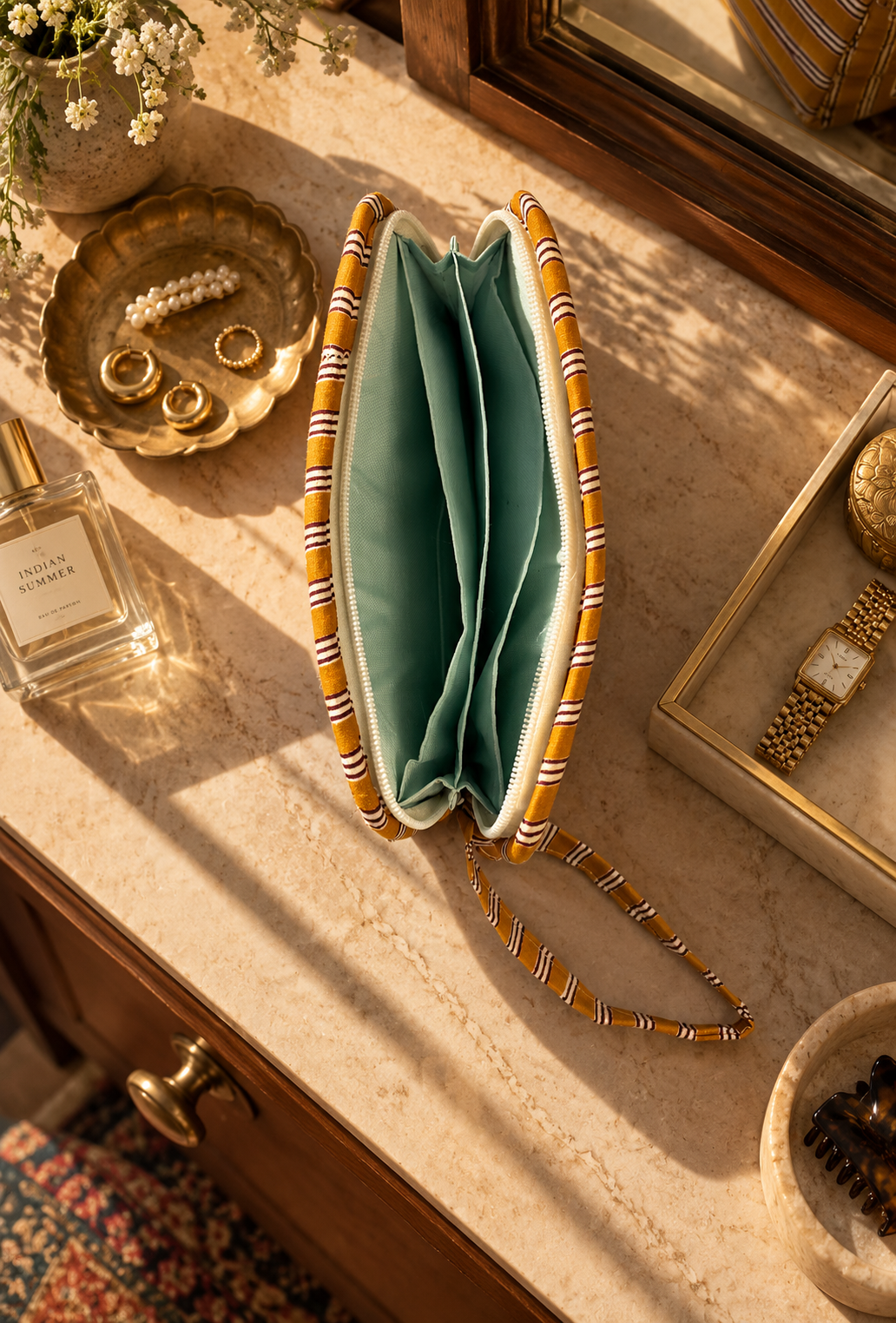 A striped fabric wallet clutch pouch with a water-resistant interior, styled on a marble vanity table with gold jewellery, a perfume bottle, and a watch nearby.