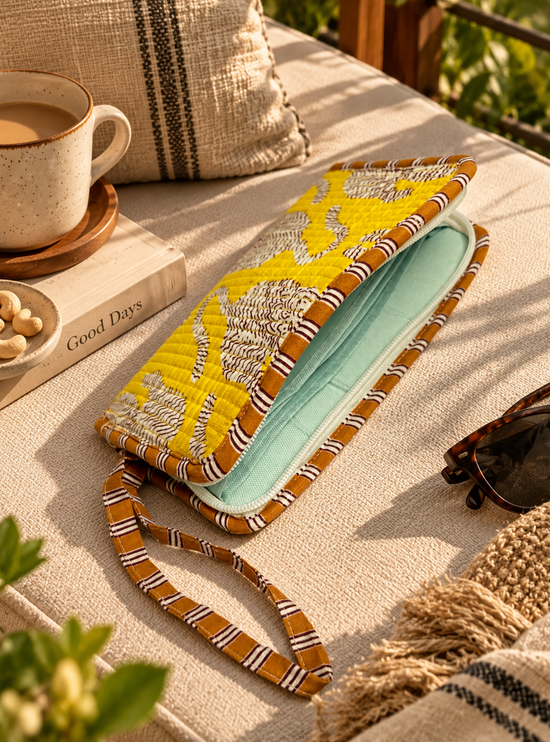 A quilted zippered clutch bag with a yellow and white floral-patterned exterior and a water-resistant interior, styled on a beige linen surface with a coffee cup, book, and sunglasses nearby.