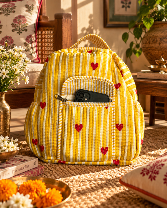 A mini backpack with yellow & white stripes with mini red hearts all over, with an open front zipper carrying a smart phone placed on a rug in front of a wooden couch. along with other Indian decorative items. 