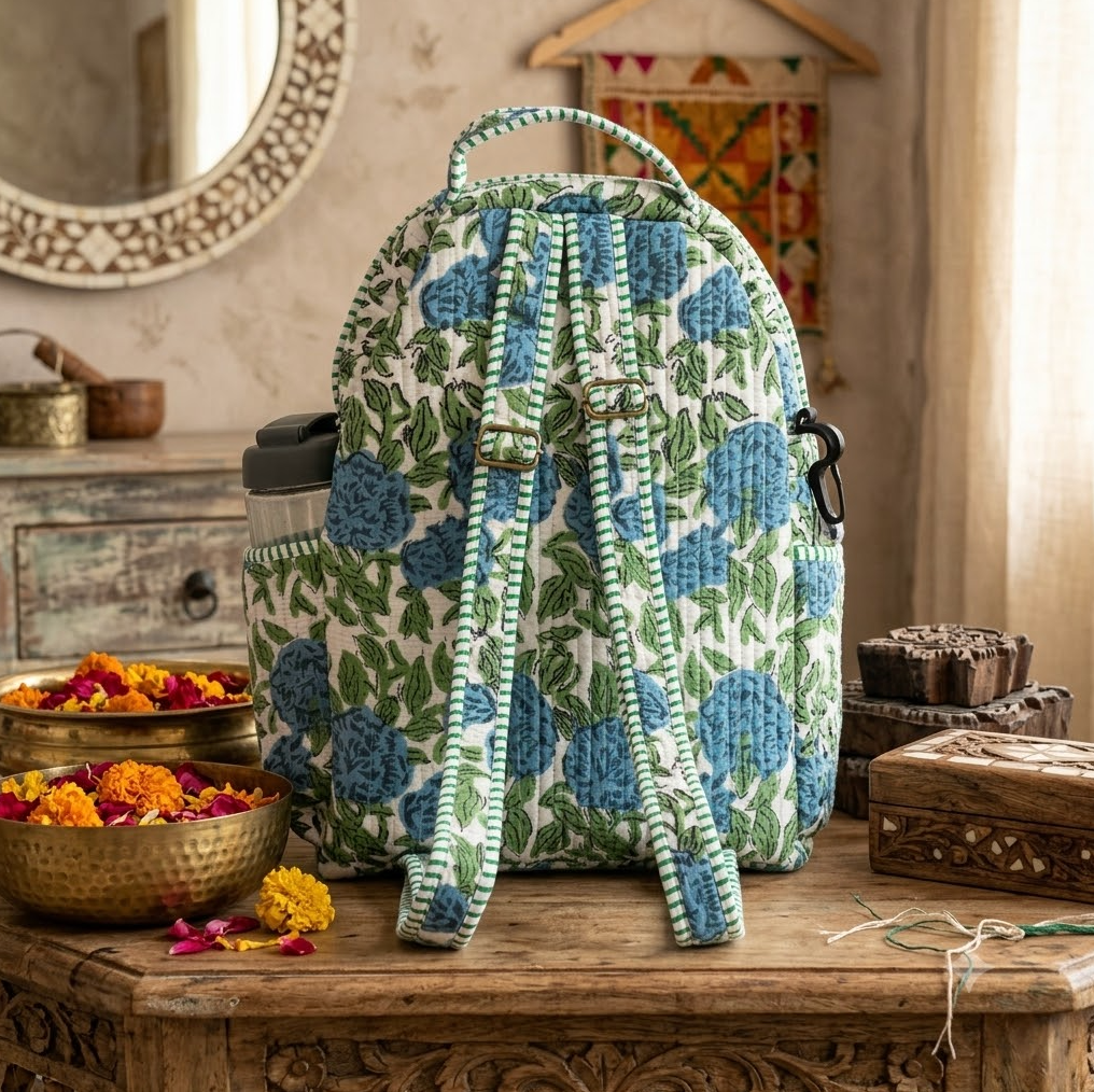 A quilted backpack with a blue and green floral block print pattern, with adjustable straps with metal adjusters styled on a rustic wooden table in a warm, bohemian room setting.