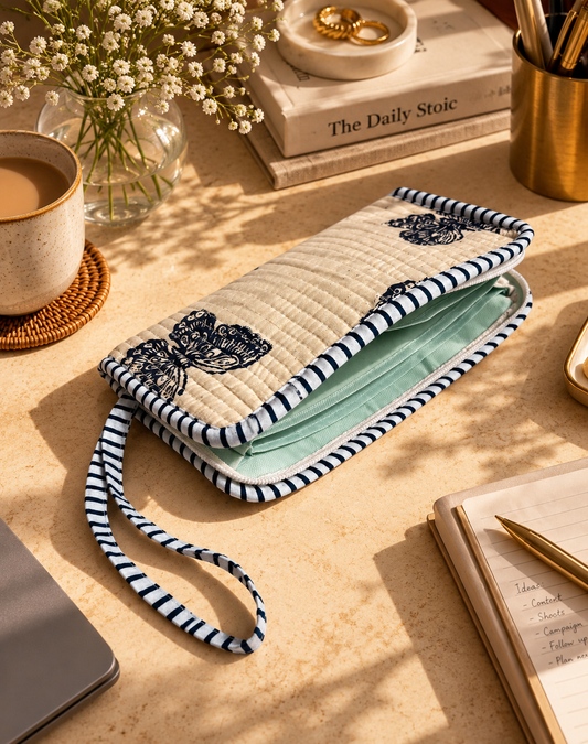 A quilted fabric clutch or wallet with a navy and white striped strap and border, featuring butterfly motifs on a white background. The wallet is shown open, revealing a light water-resistant  interior, styled on a warm beige desk with a coffee cup, books, and stationery.