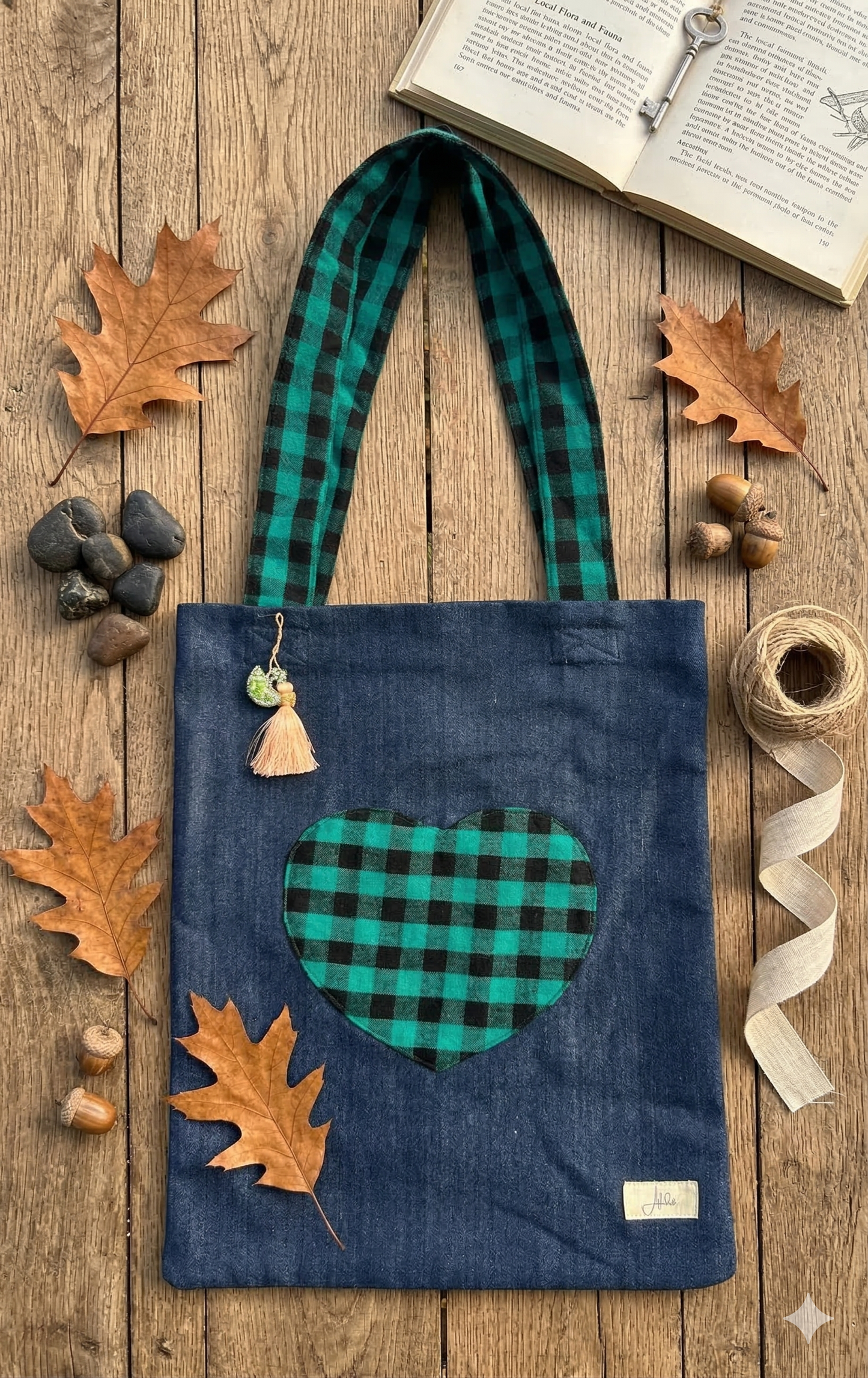 Blue Denim tote bag with green and black heart design front pocket and straps on a wooden surface with autumn leaves and rocks.