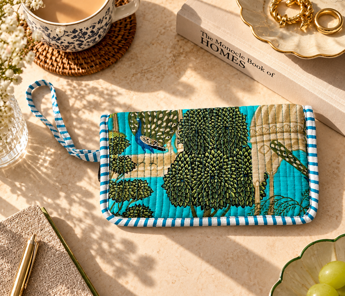 A Rajasthani print with peacock, royal courtyard and trees on a blue base quilted cotton wallet with handheld string and blue and white stripes kept on a marble table with other accessories such as a book, a cup of tea, a book and a pen. 